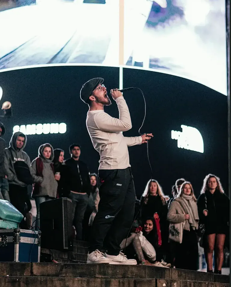 Performer singing into a microphone on steps in front of a large screen, with a crowd watching