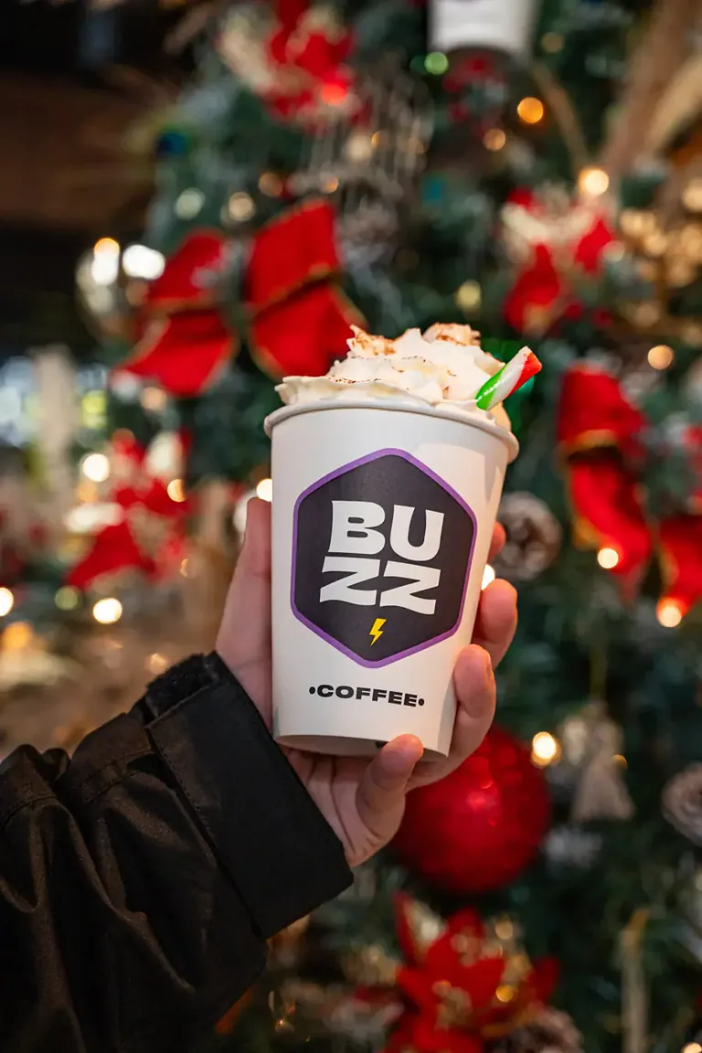 Hand holding a Buzz Coffee cup topped with whipped cream in front of a Christmas tree