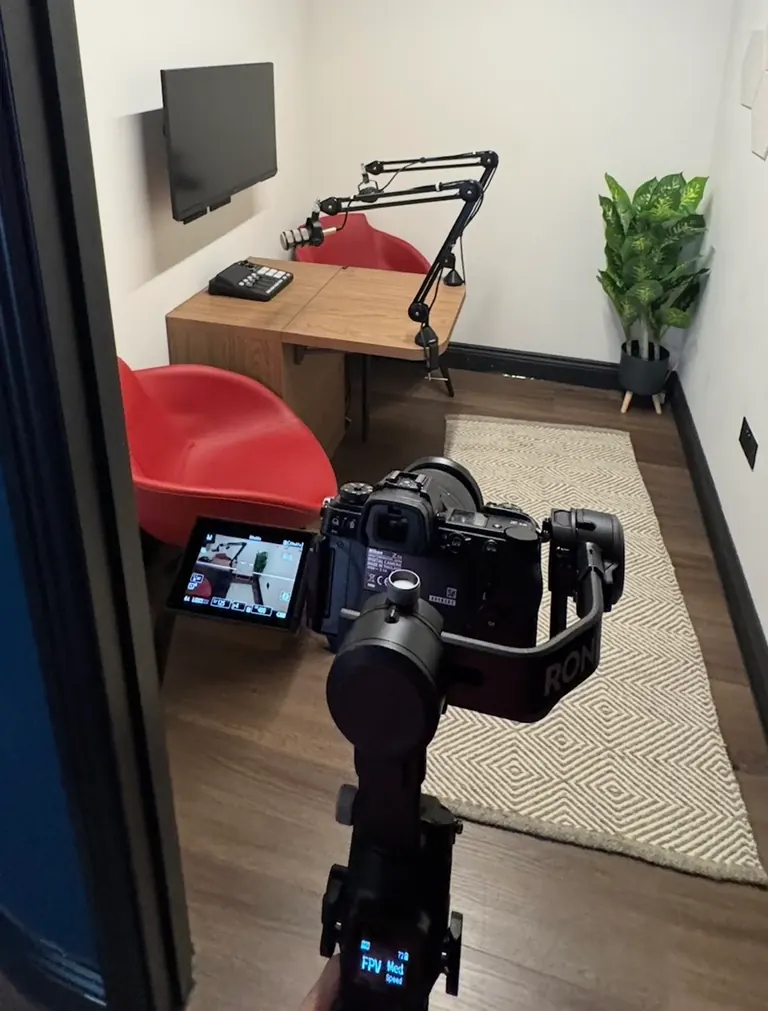 Camera on a handheld gimbal filming in a small podcast studio with microphones on boom arms