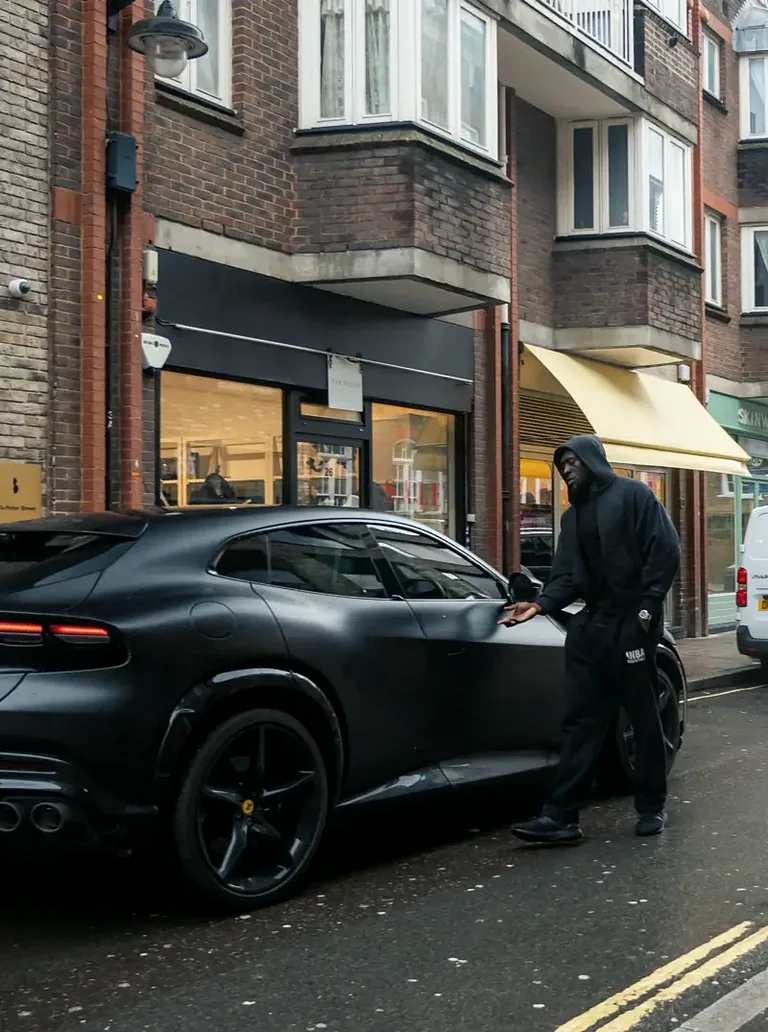 Person in a dark hoodie opening the door of a black sports car on a London street